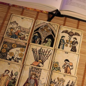 May include: A collection of nine tarot cards arranged in a grid, featuring various illustrations including figures, swords, and coins. Each card displays Roman numerals and detailed line art with muted color accents, resting on a bamboo mat surface.