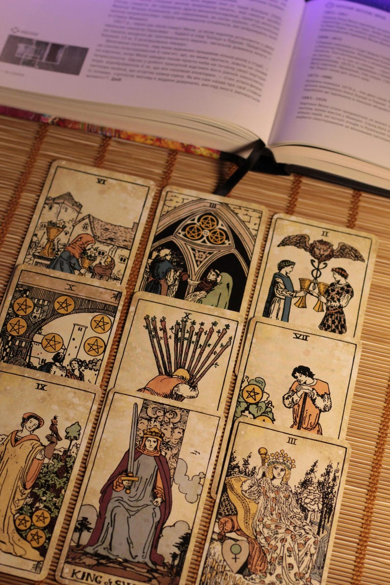 May include: A collection of nine tarot cards arranged in a grid, featuring various illustrations including figures, swords, and coins. Each card displays Roman numerals and detailed line art with muted color accents, resting on a bamboo mat surface.