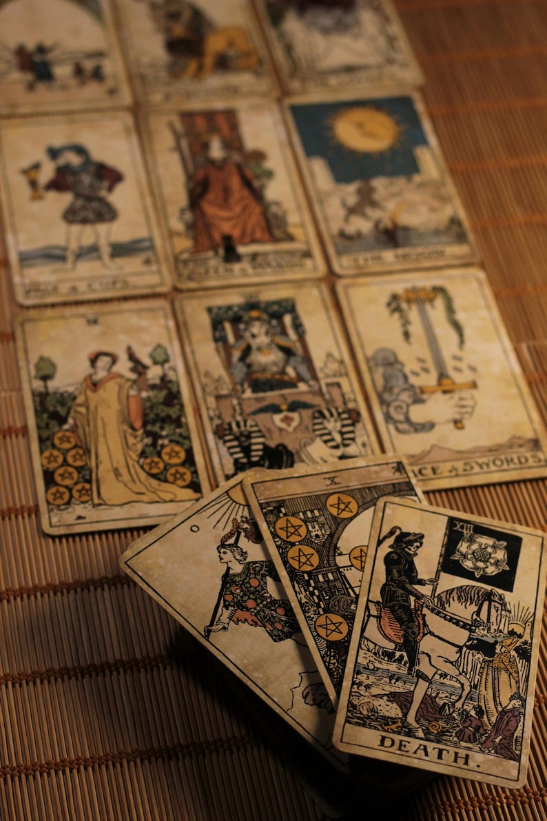 Puede incluir: Una colecci&oacute;n de cartas de tarot de estilo vintage dispuestas en una cuadr&iacute;cula y en abanico. Las cartas presentan ilustraciones detalladas, incluyendo la carta de la Muerte, el As de Espadas y la Luna, en tonos envejecidos sobre cartulina.