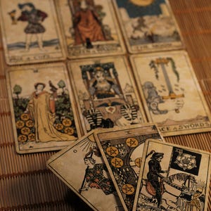 May include: A collection of vintage-style tarot cards arranged in a grid and a fan pattern. The cards feature detailed illustrations including the Death card, the Ace of Swords, and the Moon, all rendered in muted, aged tones on rectangular card stock.