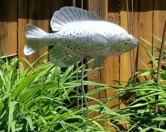 May include: A metal garden sculpture of a silver fish with a detailed design. The fish is mounted on a metal rod and is surrounded by green grass.