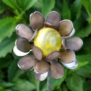 May include: A metal flower sculpture with a yellow and white marble in the center. The flower has 12 petals that are a dark brown color with a silver metallic finish.