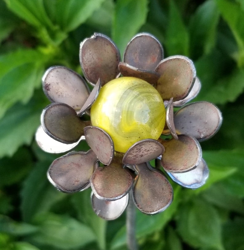 May include: A metal flower sculpture with a yellow and white marble in the center. The flower has 12 petals that are a dark brown color with a silver metallic finish.