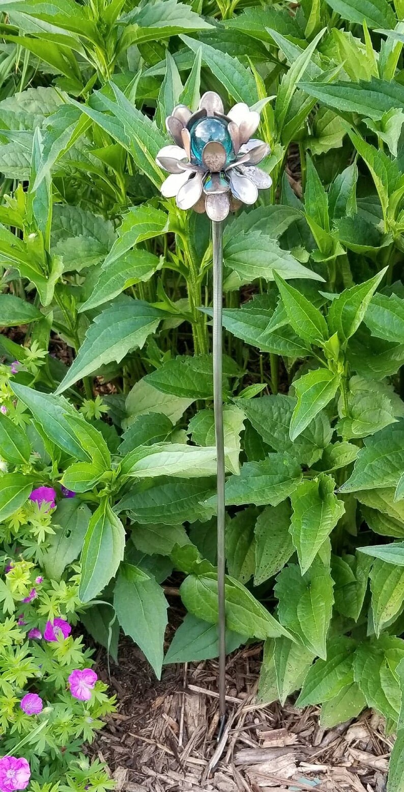 May include: A metal flower sculpture with a blue glass center, standing in a bed of green plants.