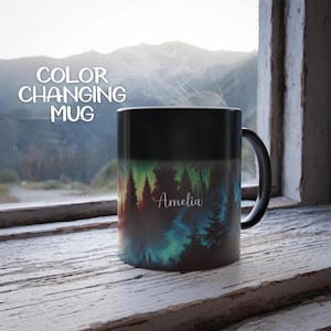 May include: Black color changing mug with a colorful forest scene and the name Amelia printed on it. The mug is sitting on a wooden surface.