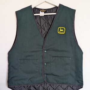 May include: Dark green vest with two front pockets and a black quilted lining. The vest has a V-neck and a small yellow and green John Deere logo patch. It is buttoned up and hanging on a white hanger.