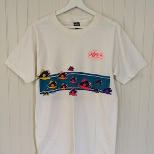 May include: White short-sleeved t-shirt with a graphic of colorful fish swimming in a blue and green stripe. The shirt has a red and white logo that says "Mossy Oak" with the words "The Original" and "1986" in smaller font.