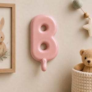 May include: A glossy, light pink wall hook shaped like the uppercase letter B. The hook features a smooth, rounded surface with two circular openings in the centre of the letter and a curved protrusion at the bottom for hanging items.