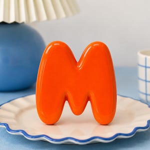 May include: A glossy, bright orange, three-dimensional letter M with a smooth, rounded, bubble-like shape. The letter stands upright in the center of a white ceramic plate with a scalloped edge and a thin blue rim.