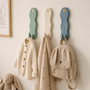 May include: Three vertical wall hooks with wavy edges in sage green, cream, and dusty blue. Each hook features a gold-toned screw at the top. A cream knit cardigan, a patterned beige muslin cloth, and a beige bunny-shaped backpack hang from them.
