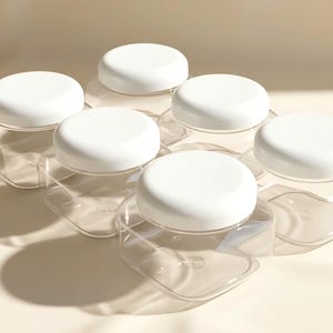 May include: Six clear, hexagonal-shaped jars with white, round lids are arranged in a cluster. The jars are empty and transparent, with the lids providing a contrasting white color. The jars are sitting on a light-colored surface.