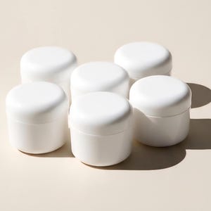 May include: Six white, round cosmetic jars with lids are arranged on a light beige surface. The jars are made of a smooth, opaque material and are evenly spaced. The lids are also white and fit snugly on top of the jars.