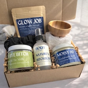 May include: A skincare gift set in a cardboard box containing a Glow Job face mask pouch, a Bye Bitch cleansing balm jar, a Nature's Bitch Slap spray bottle, a Moist AF face cream jar, a wooden bowl, a white washcloth, and a white brush.
