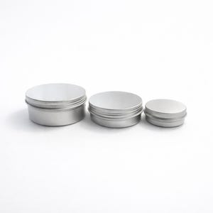 May include: Three silver-colored, round metal tins of varying sizes. Each tin has a lid and a ridged edge. The tins are arranged from largest to smallest, suggesting a set for storage or packaging.