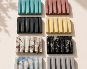 May include: Eight rectangular soap dishes arranged in two columns. The dishes feature vertical drainage grooves and come in various finishes: solid teal, dusty rose, black, yellow, white with brown marbling, black with gold marbling, blue and white swirl, and gray.