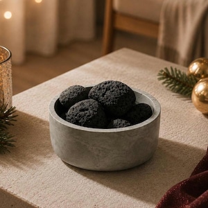 May include: A round, light gray concrete bowl filled with several dark black, porous lava stones. The bowl has a smooth, matte finish and a simple, cylindrical shape, containing the textured, irregular volcanic rocks within its basin.