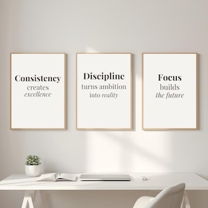Op de afbeelding: Drie ingelijste prints met motiverende citaten: "Consistentie creëert excellentie", "Discipline zet ambitie om in realiteit" en "Focus bouwt de toekomst". De prints hebben een witte achtergrond met zwarte tekst en zijn in lichtbruine lijsten.