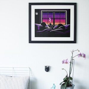 May include: A framed art print depicts two figures in bed, smoking, with a sunset view through a window. The artwork features a black frame, a purple and orange sky, and a cannabis leaf symbol. The print is mounted on a white wall.