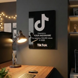 Könnte beinhalten: Eine schwarz-weiße TikTok-Auszeichnungstafel mit dem TikTok-Logo und dem Text "Presented to: YOUR NICKNAME For passing 100,000 views in 2026". Die Tafel hängt an einer grauen Wand über einem Holzschreibtisch.