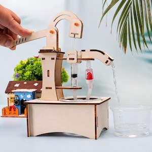 May include: A wooden hydraulic water pump model kit featuring a hand-operated lever, a clear plastic syringe, and tubing. The structure is made of light-colored plywood with dark edges, designed to pump water from a clear plastic cup into a basin.