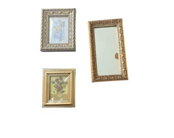 May include: Three ornate, gold-toned picture frames against a white background. One frame contains a floral print, another a mirror, and the third a painting of sunflowers in a vase. The frames have detailed, decorative edges.