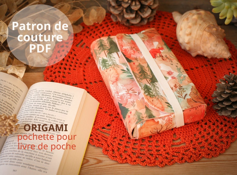 May include: A PDF sewing pattern for an origami book clutch. The clutch is made of fabric with a floral print and has a white ribbon closure. The pattern is titled "Origami Pochette pour Livre de Poche".