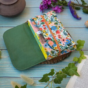 May include: A green fabric book cover with a floral patterned book inside. The book cover is closed and the book is visible through the open flap. The book cover is sitting on a wooden surface.