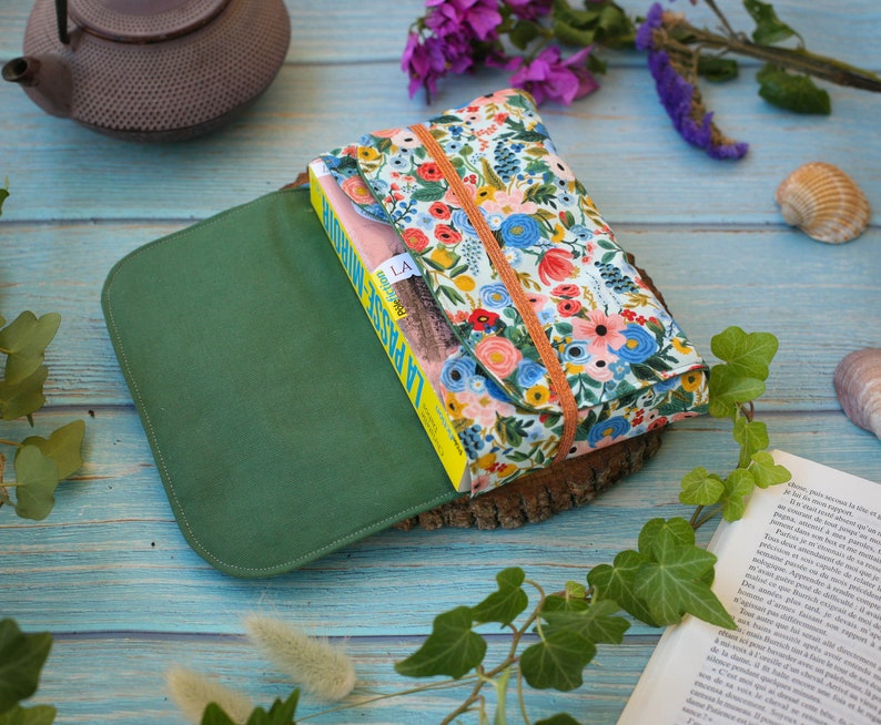 May include: A green fabric book cover with a floral patterned book inside. The book cover is closed and the book is visible through the open flap. The book cover is sitting on a wooden surface.
