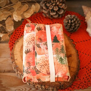 May include: A small, rectangular, fabric pouch with a white strap. The pouch is decorated with a watercolor-style print of a forest scene with trees, deer, and leaves in shades of orange, pink, and green.