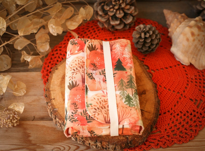 May include: A small, rectangular, fabric pouch with a white strap. The pouch is decorated with a watercolor-style print of a forest scene with trees, deer, and leaves in shades of orange, pink, and green.