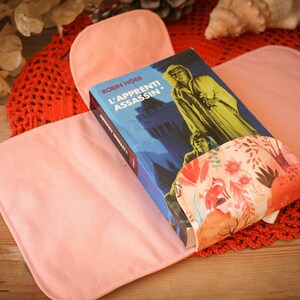 May include: A pink fabric book cover with a floral pattern on the inside. The cover is holding a book titled "L'Apprenti Assassin" by Robin Hobb.