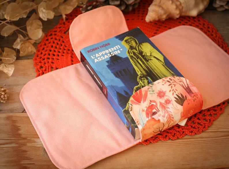 May include: A pink fabric book cover with a floral pattern on the inside. The cover is holding a book titled "L'Apprenti Assassin" by Robin Hobb.