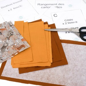 May include: A sewing project with fabric pieces, a pair of scissors, and instructions for a fabric pouch. The fabric pieces are in shades of brown and orange, and the instructions are written in French.