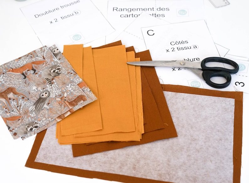 May include: A sewing project with fabric pieces, a pair of scissors, and instructions for a fabric pouch. The fabric pieces are in shades of brown and orange, and the instructions are written in French.