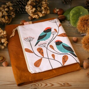 May include: A white zippered pouch with a brown suede cover. The pouch features a design of two blue and turquoise birds with red accents perched on branches with orange leaves.