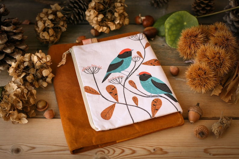 May include: A white zippered pouch with a brown suede cover. The pouch features a design of two blue and turquoise birds with red accents perched on branches with orange leaves.