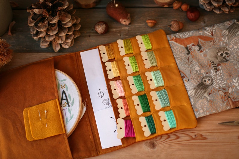 May include: A mustard yellow felt embroidery kit with a wooden organizer for floss. The kit includes a hoop with a floral design and the letter "A" embroidered on it, a printed pattern, and a fabric with a woodland animal print.