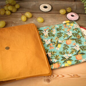May include: A mustard yellow fabric clutch with a zipper closure. The clutch is open to reveal a turquoise fabric lining with a floral pattern of white flowers, green leaves, and yellow lemons.