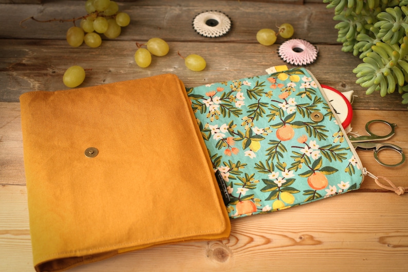 May include: A mustard yellow fabric clutch with a zipper closure. The clutch is open to reveal a turquoise fabric lining with a floral pattern of white flowers, green leaves, and yellow lemons.