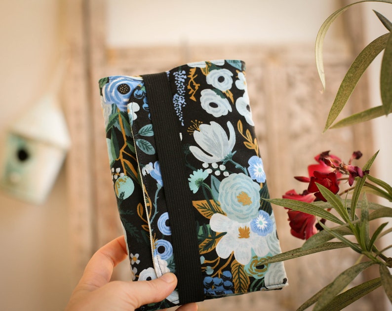 May include: A black fabric pouch with a floral pattern in shades of blue, green, and yellow. The pouch has a black strap and a zipper closure.