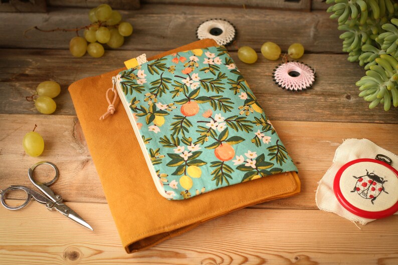 May include: A mustard yellow fabric notebook cover with a zippered pouch. The pouch is made of a light blue fabric with a floral pattern of white flowers, green leaves, and orange citrus fruit.