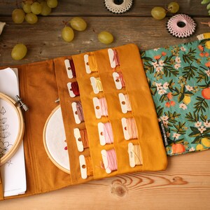 May include: A mustard yellow fabric roll-up case with multiple compartments for storing embroidery floss. The case has a floral print fabric flap with lemons and flowers. The case is open to reveal the compartments and a small hoop.
