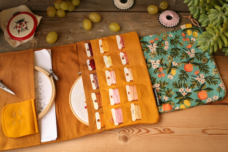 May include: A mustard yellow fabric roll-up case with multiple compartments for storing embroidery floss. The case has a floral print fabric flap with lemons and flowers. The case is open to reveal the compartments and a small hoop.