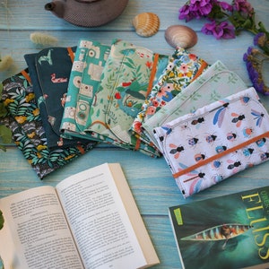 May include: A collection of six colorful fabric book covers with various patterns, including floral, geometric, and nature-inspired designs. The covers are arranged on a blue wooden surface with a book and a few decorative items.