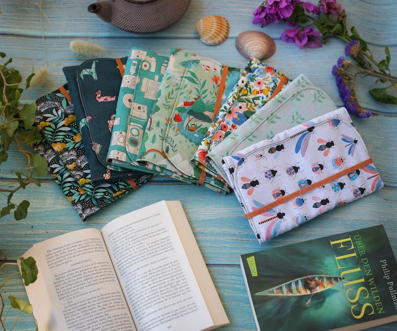 May include: A collection of six colorful fabric book covers with various patterns, including floral, geometric, and nature-inspired designs. The covers are arranged on a blue wooden surface with a book and a few decorative items.