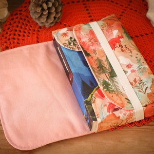 May include: A pink fabric book cover with a floral pattern and a white elastic band. The cover is open to reveal a blue book with a white title.