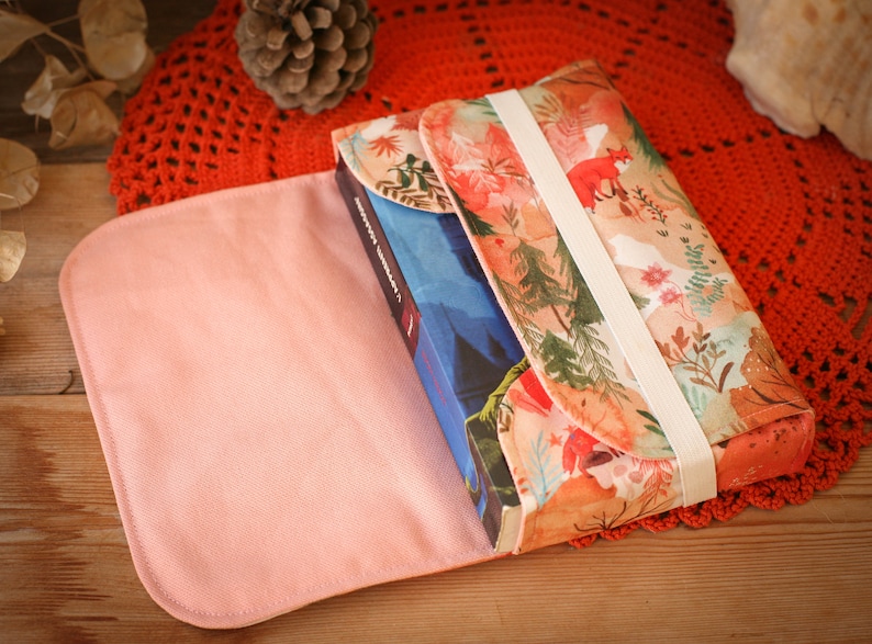 May include: A pink fabric book cover with a floral pattern and a white elastic band. The cover is open to reveal a blue book with a white title.
