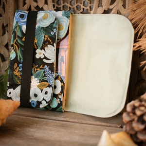 May include: A black fabric book cover with a floral pattern in shades of blue, white, and yellow. The cover has a black elastic band to keep it closed. The cover is open to reveal a plain beige fabric lining.