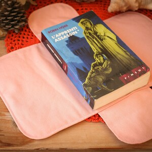 May include: A pink fabric book cover with a book titled "L'APPRENTI ASSASSIN." by Robin Hobb. The book cover features a blue background with a castle and a person in a hooded robe.
