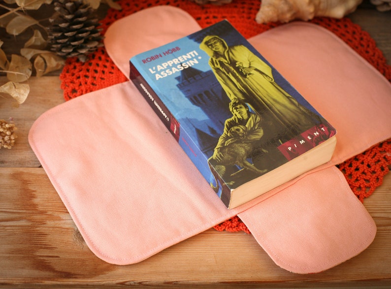 May include: A pink fabric book cover with a book titled "L'APPRENTI ASSASSIN." by Robin Hobb. The book cover features a blue background with a castle and a person in a hooded robe.